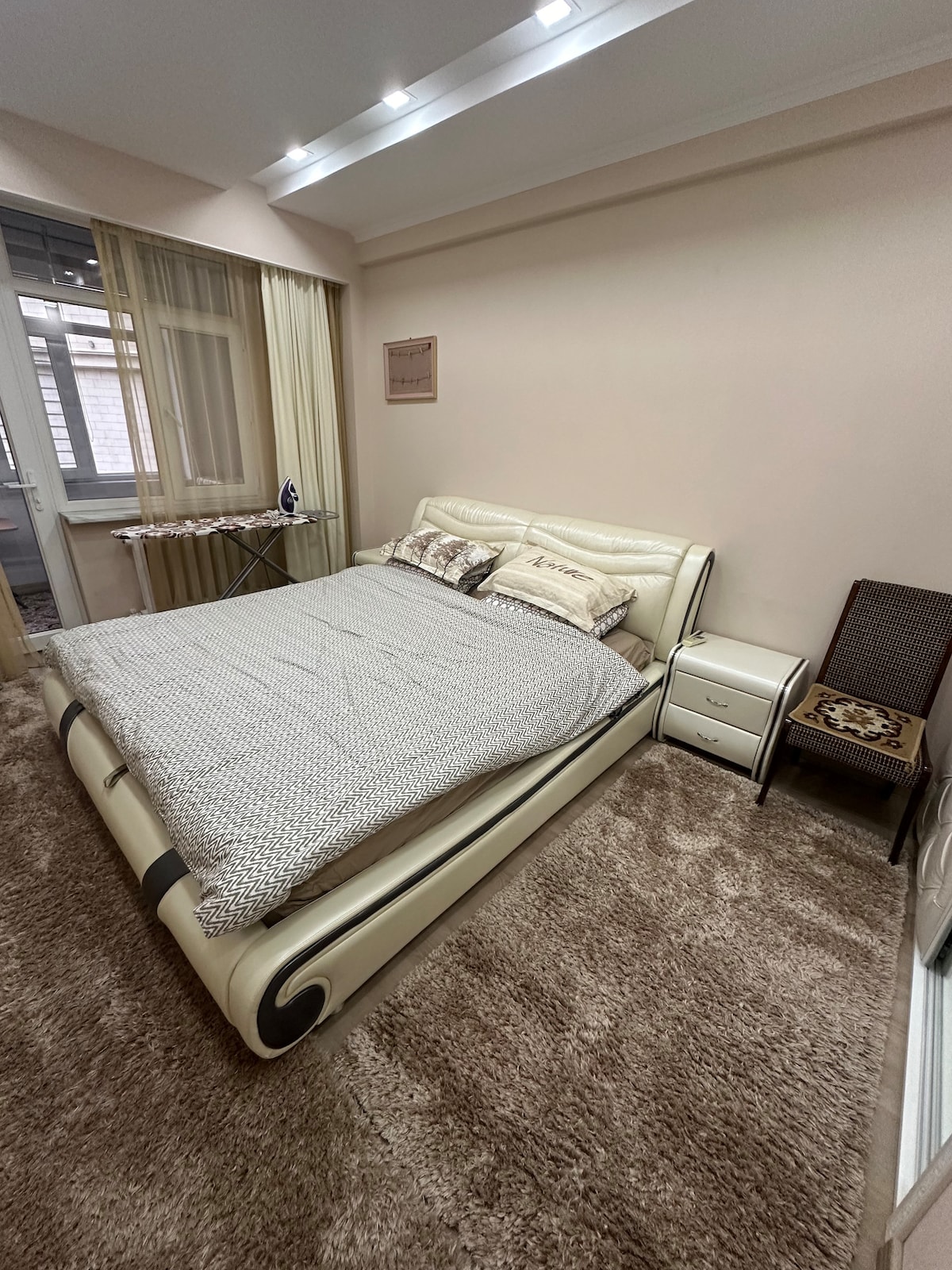 A well-lit bedroom features a spacious bed dressed with a soft, textured blanket. A bedside table stands next to the bed, accompanied by a chair with a patterned cushion. Light beige walls and curtains contribute to a bright, calming environment.