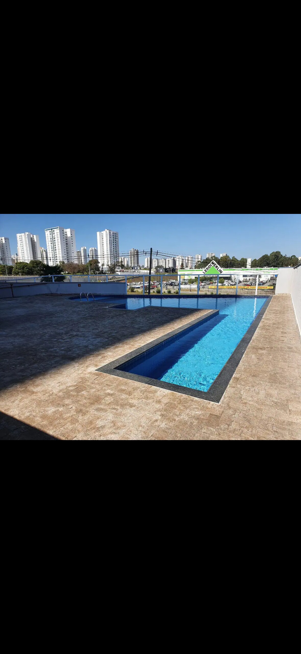 A modern swimming pool is visible, featuring a long, narrow design filled with clear blue water. Sunlight reflects off the surface, and tall buildings in the background provide a cityscape view. A spacious pool deck surrounds the area, enhancing the overall outdoor ambiance.