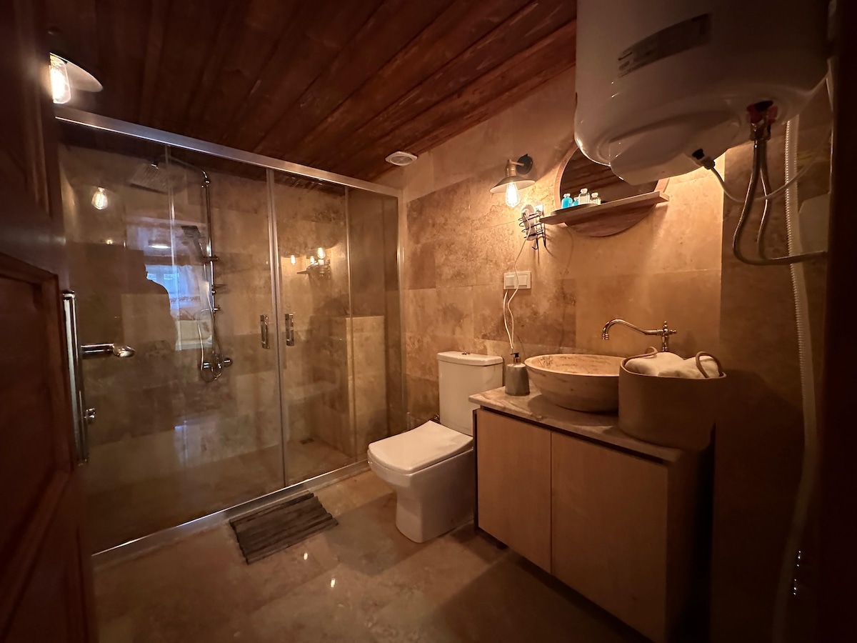 The bathroom features natural stone walls and warm wooden accents. A spacious glass shower is equipped with multiple shower heads. A modern toilet is positioned next to a wooden vanity, which contains a round stone sink and neatly arranged towels.