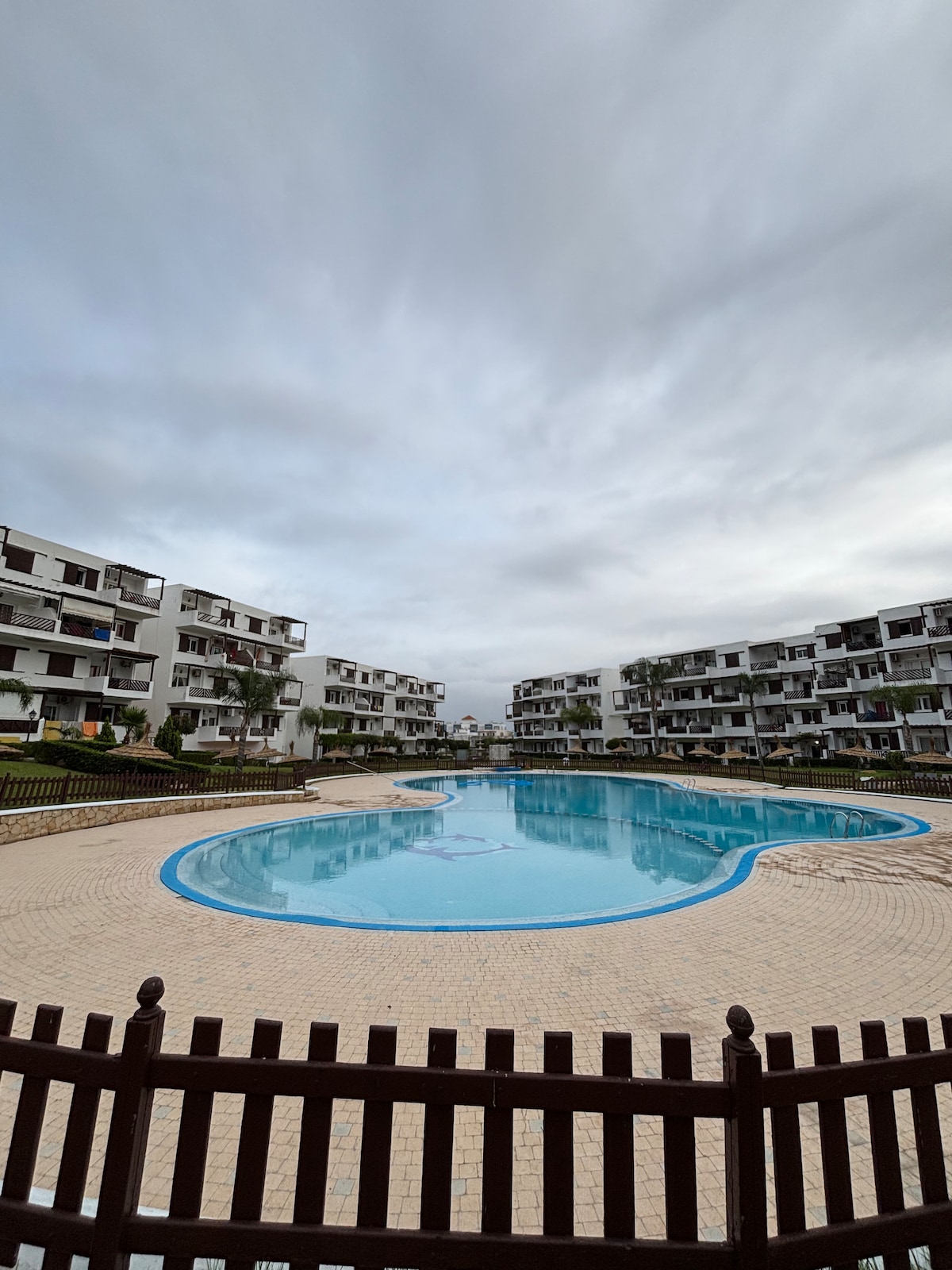 A spacious outdoor area is presented, featuring a large swimming pool surrounded by neatly arranged lounge chairs. The pool is bordered by a light-colored walkway and low wooden fence. Nearby, several multi-story buildings are visible, creating a cozy residential atmosphere.