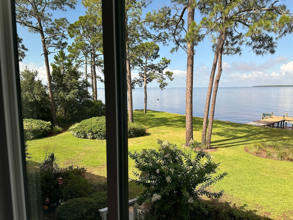 A serene view of Chotawhatchee Bay is presented, framed by tall pine trees and lush greenery. The calm waters reflect the blue sky, creating a peaceful atmosphere. A grassy area extends towards the water, leading to a wooden pier in the distance.