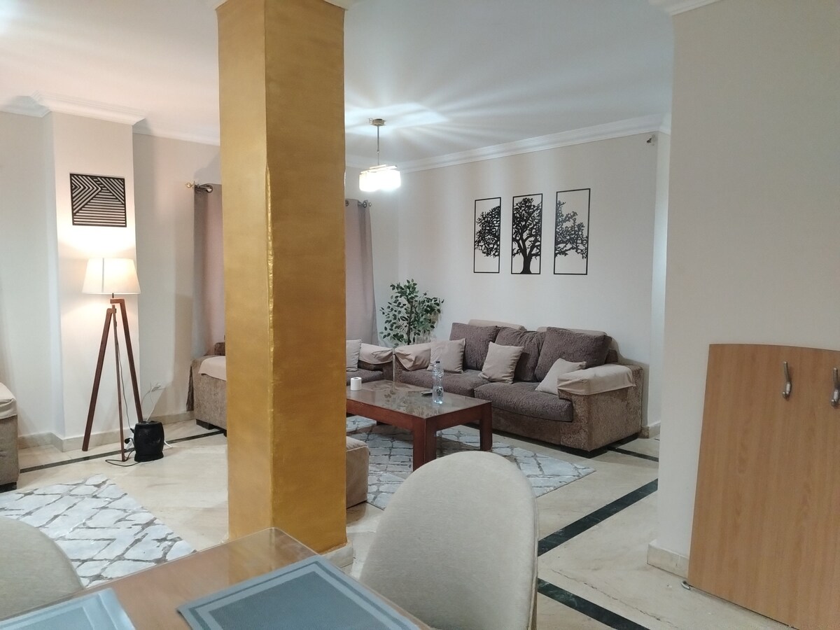 A welcoming living area features soft, neutral-toned sofas arranged around a wooden coffee table. A tall lamp provides soft lighting beside a decorative wall with three framed tree artworks. Light curtains frame the windows, and a potted plant adds a touch of greenery.