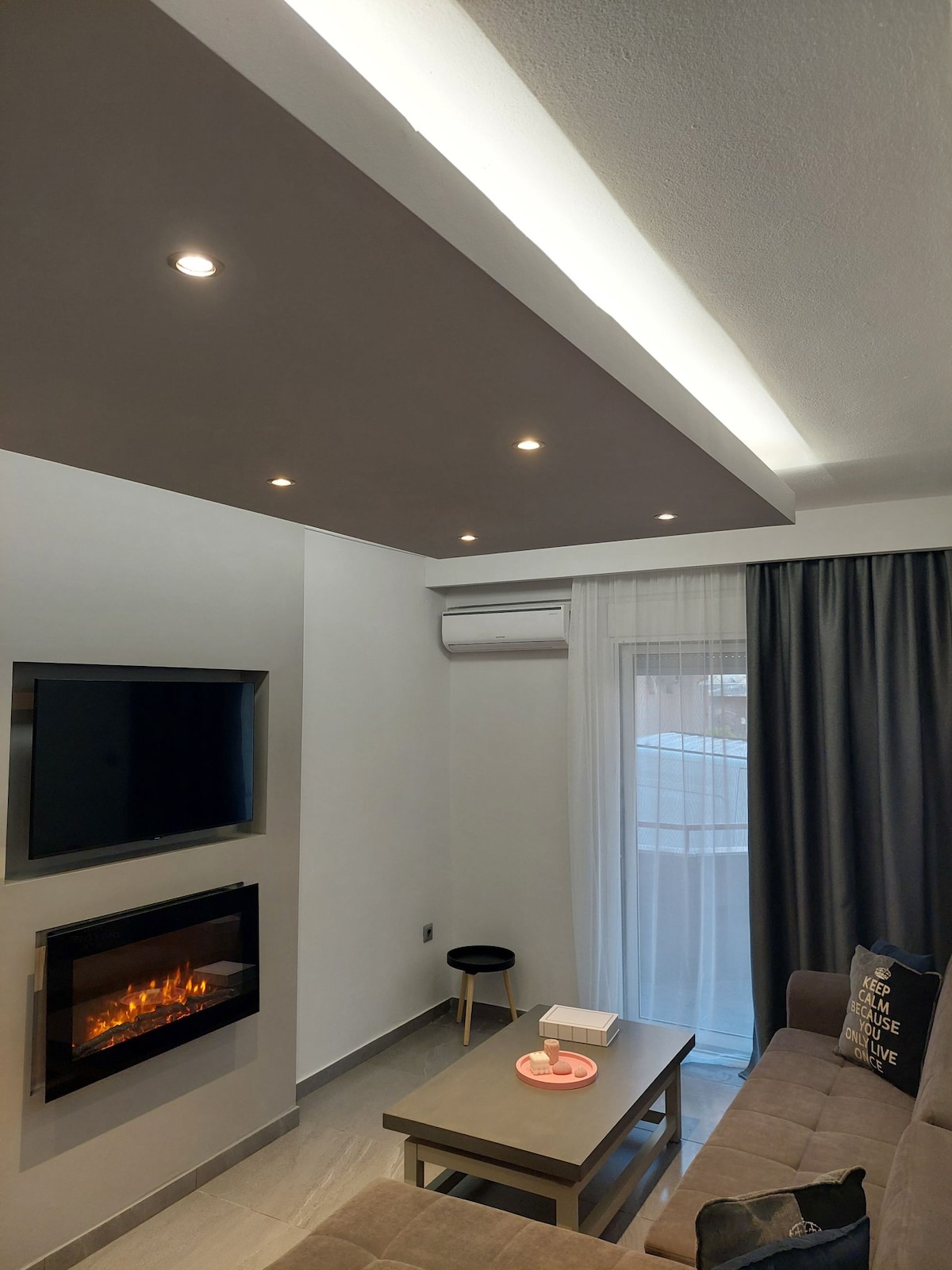 A modern living area features a wall-mounted television above a sleek entertainment unit. A cozy electric fireplace is set into the wall, adding warmth. Soft curtains frame a window, allowing natural light to enter, while a small, minimalistic stool and a coffee table complete the space.