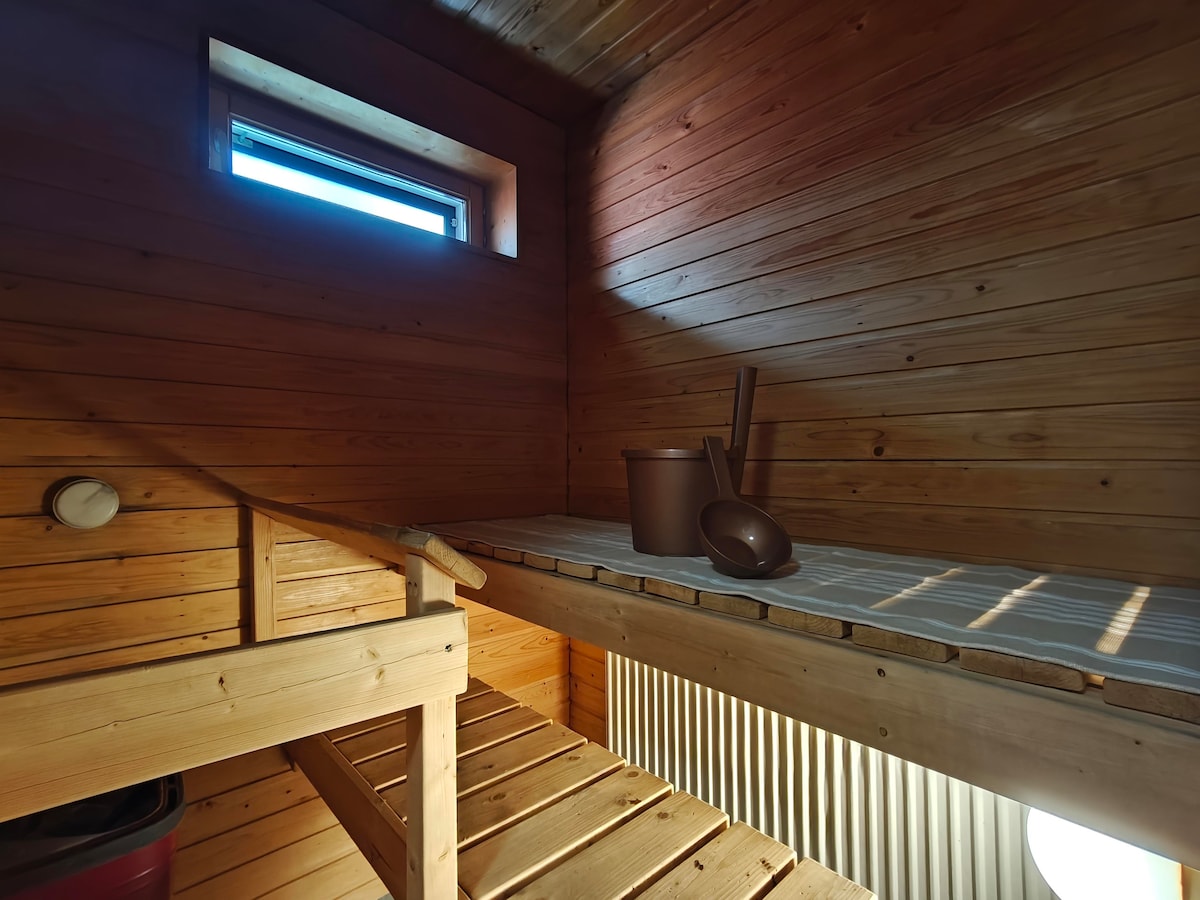 A wooden sauna interior is presented, featuring a small bench with a soft mat and a bucket alongside a scoop. Natural light filters through a small window, illuminating the warm wooden walls and creating a calming atmosphere.