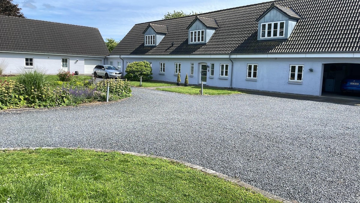 A spacious gravel driveway leads to a charming two-story building, featuring multiple gabled windows. Lush green grass borders the driveway, while well-maintained flower beds add color to the landscape. A car is parked nearby, and additional buildings are visible in the background.
