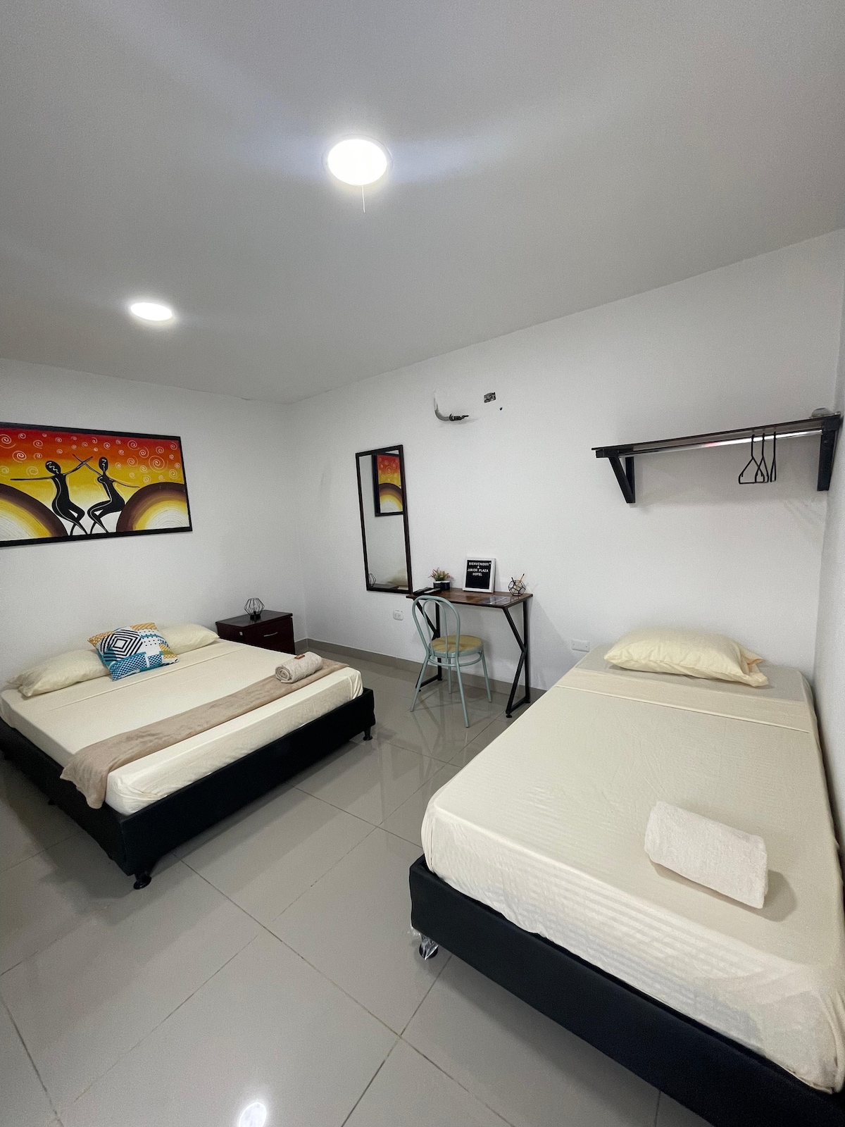 The room includes a double bed and a single bed, both with light bedding. A small desk and chair are positioned near a wall-mounted shelf. Artwork is displayed above the double bed, and soft lighting illuminates the neutral-toned walls and tiled flooring.