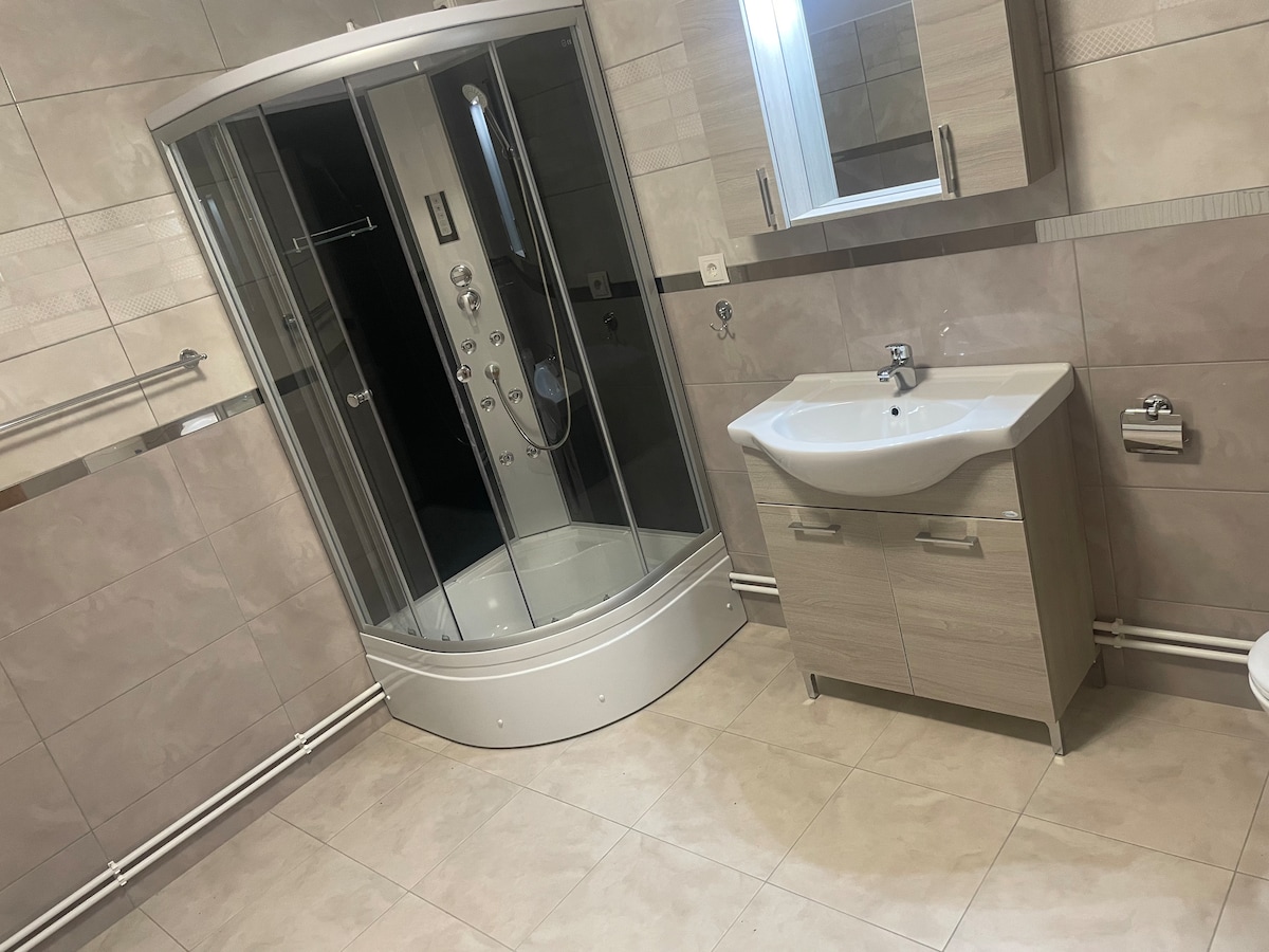 The bathroom features a modern shower enclosure with multiple controls and a spacious vanity unit. Light-colored tiles line the walls and floor, creating a bright and clean appearance. A mirror is mounted above the sink, complementing the contemporary design.