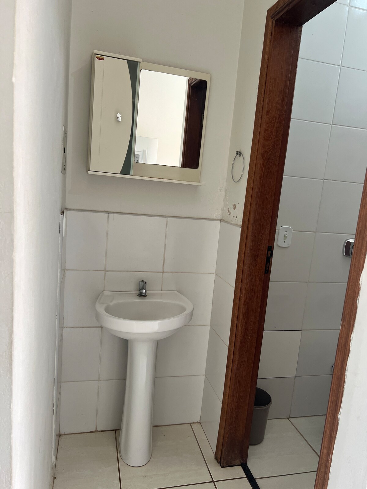A clean bathroom is visible, featuring a compact sink with a mirror above it. White tiles cover the walls, and a small waste bin is placed by the door. A shower area is located behind a wooden door.