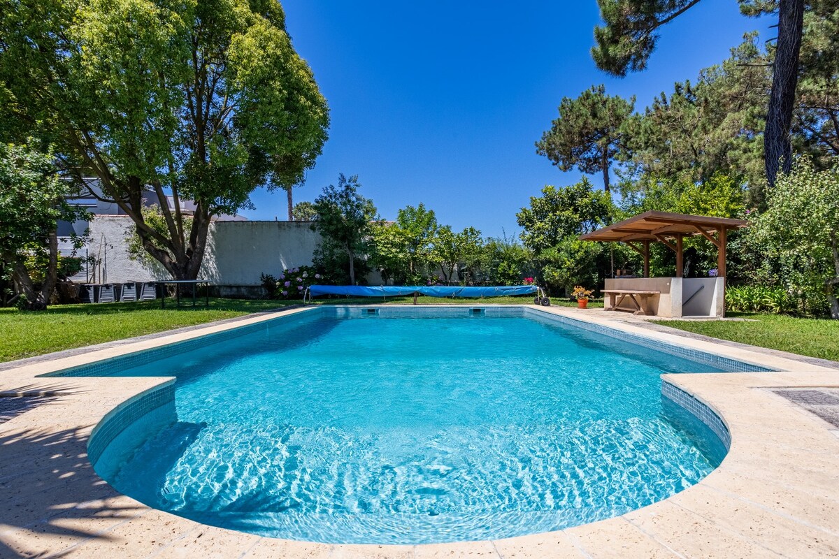 A spacious swimming pool is surrounded by lush greenery and trees. Natural grass borders the pool area, while a shaded pavilion is visible in the background, featuring a seating area. Clear blue skies enhance the inviting atmosphere of the outdoor space.