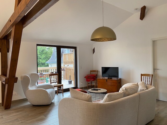 A spacious living area features a light-colored sofa and two armchairs arranged around a circular coffee table. Large windows bring in natural light and offer views of an outdoor space, while wooden beams add character to the high ceiling.