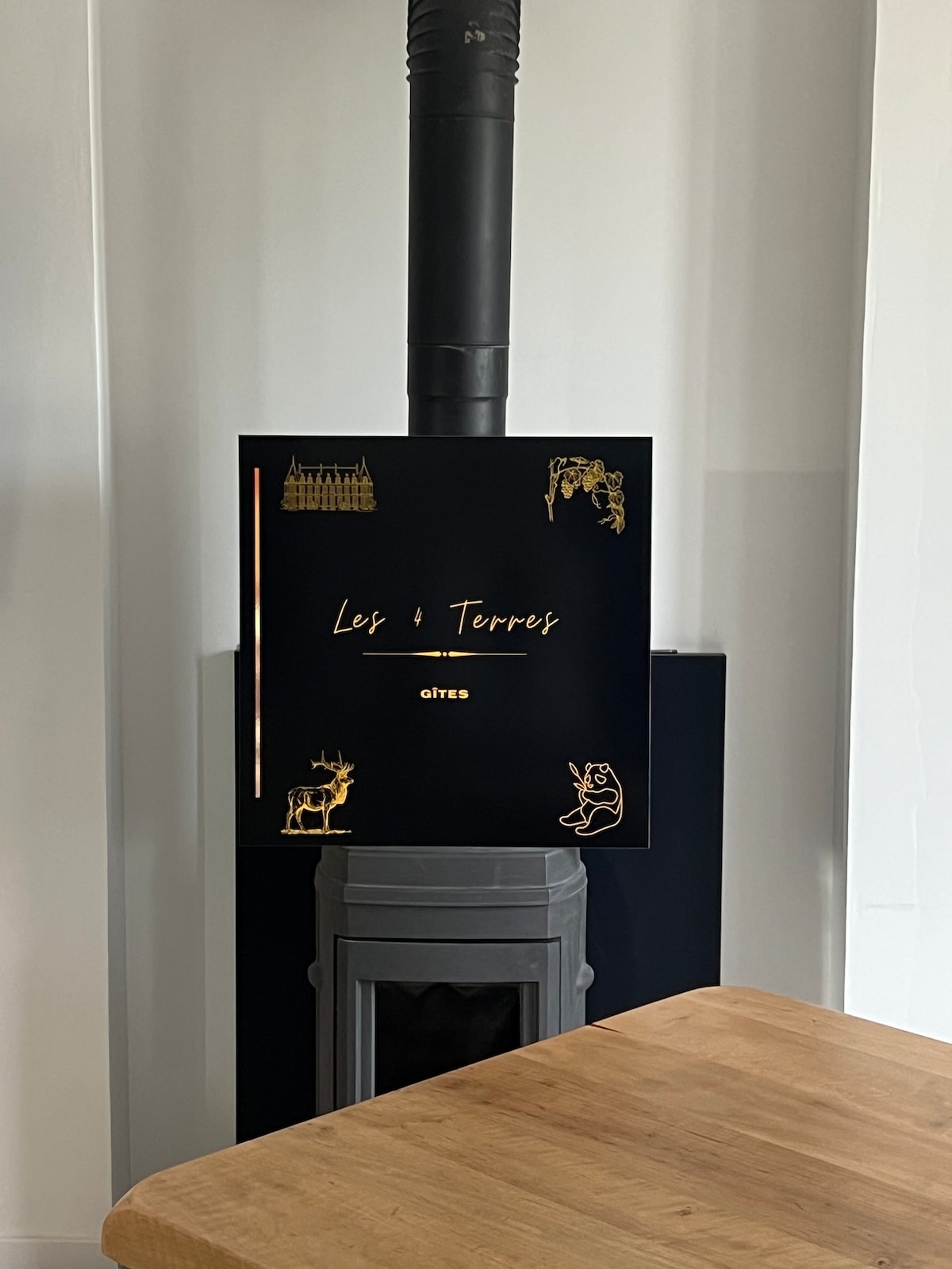 A contemporary wood-burning stove is positioned against a sleek wall, featuring an artistic sign that reads 'Les 8 Terres.' Decorative gold illustrations adorn the sign, adding a unique touch to the minimalist interior space.