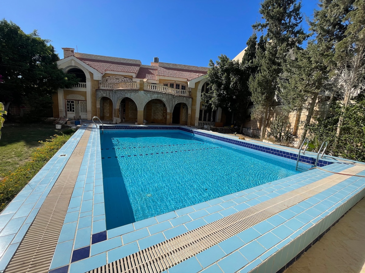 A clear blue pool is surrounded by a spacious patio with light-colored tiles. Lush greenery and well-maintained trees line the area, offering natural shade. The two-story house with arched openings stands in the background, enhancing the outdoor setting.