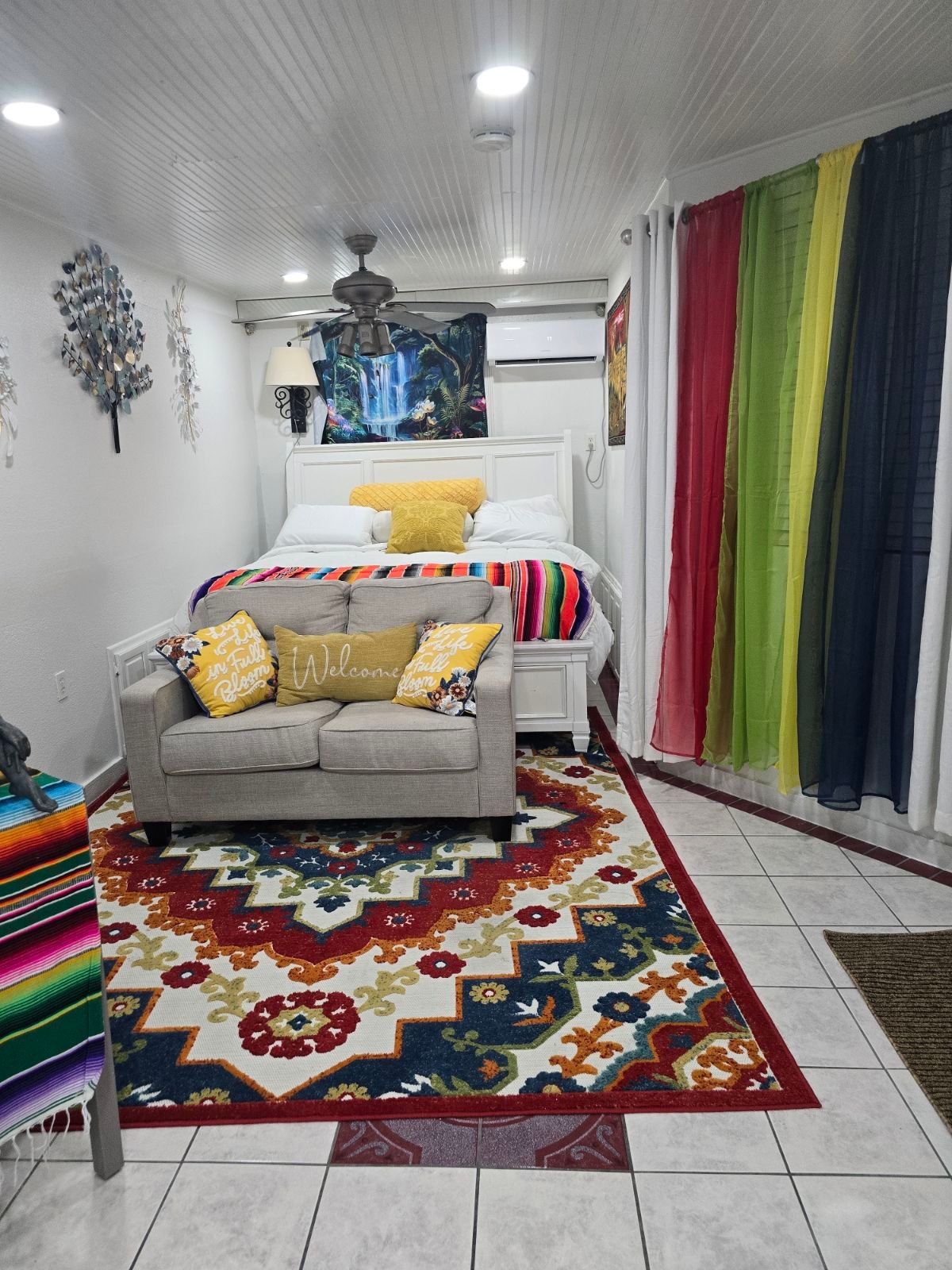 An efficiency space features a cozy bed decorated with bright yellow and patterned pillows. A comfortable seating area with a light-colored sofa sits adjacent to a colorful rug. Floor-to-ceiling curtains in various colors frame the room, enhancing the vibrant decor throughout.