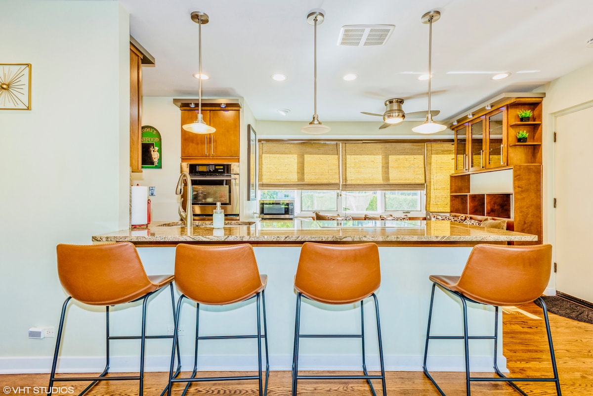 A modern kitchen area features a large granite countertop with four sleek, brown barstools positioned in front. Wooden cabinetry complements the space, while large windows allow natural light to fill the room, showcasing the organized layout and inviting atmosphere.