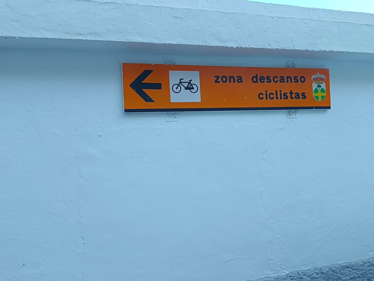 A vibrant orange sign is mounted on a white wall, indicating a cycling rest area. An icon of a bicycle is depicted to the left, directing cyclists to the zone. Text is displayed in Spanish, with the words 'zona descanso ciclistas' prominently featured.