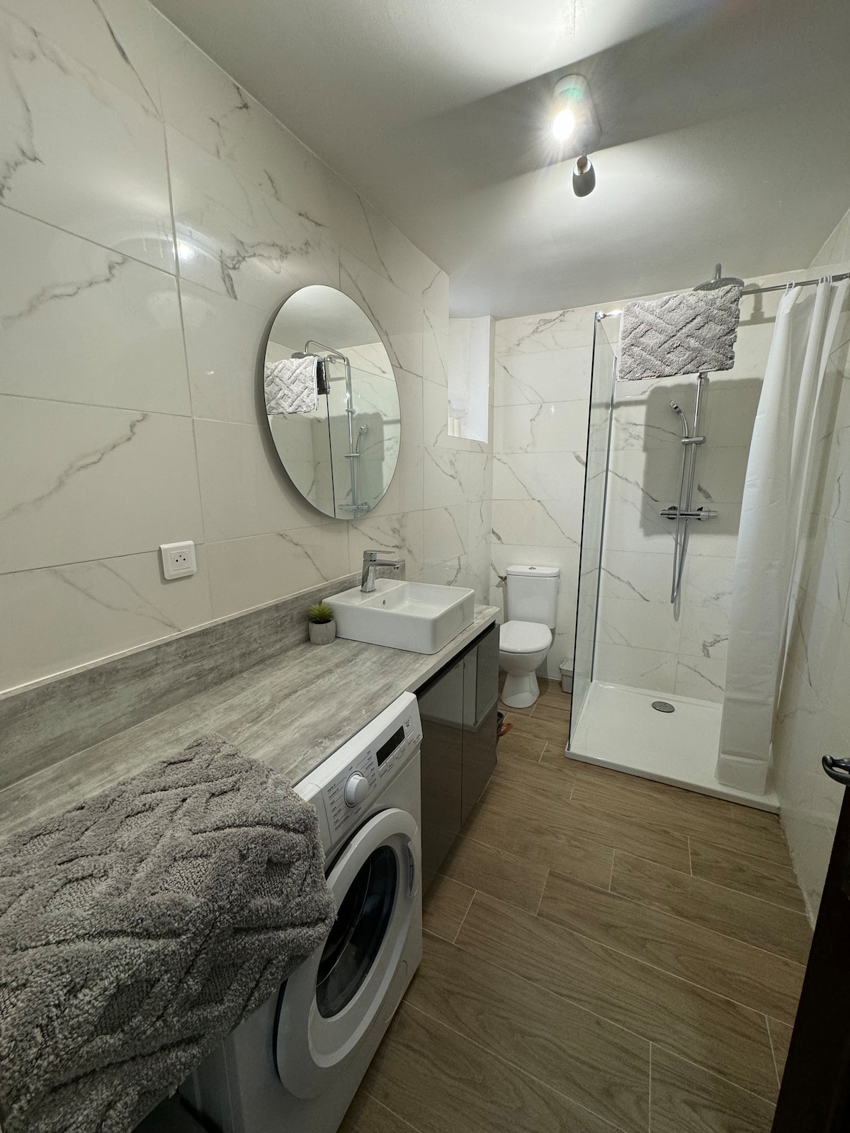 The modern bathroom features a sleek design with marble-like tiles. A round mirror is mounted above a compact sink and vanity. A washing machine is integrated into the cabinetry. A walk-in shower with a glass partition is visible, alongside a toilet, providing essential amenities.