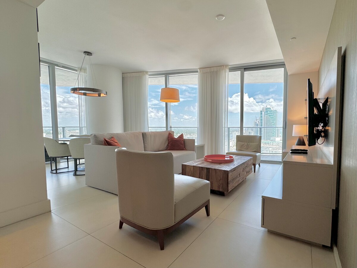 A bright and airy living area features large windows that provide ample natural light and stunning views. A modern sofa is complemented by a wooden coffee table at the center. A dining area is visible in the background, enhancing the open floor plan.
