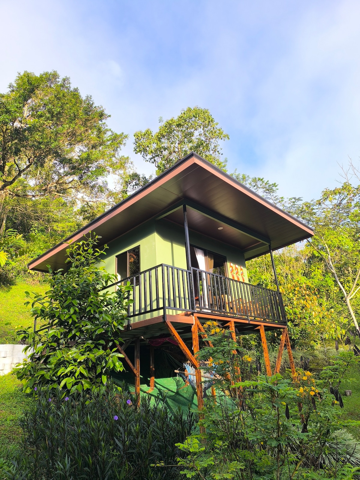 A quaint cabin is elevated on stilts, surrounded by lush greenery and vibrant plants. The structure features a simple design with a sloped roof and a small balcony, offering a peaceful retreat in nature.