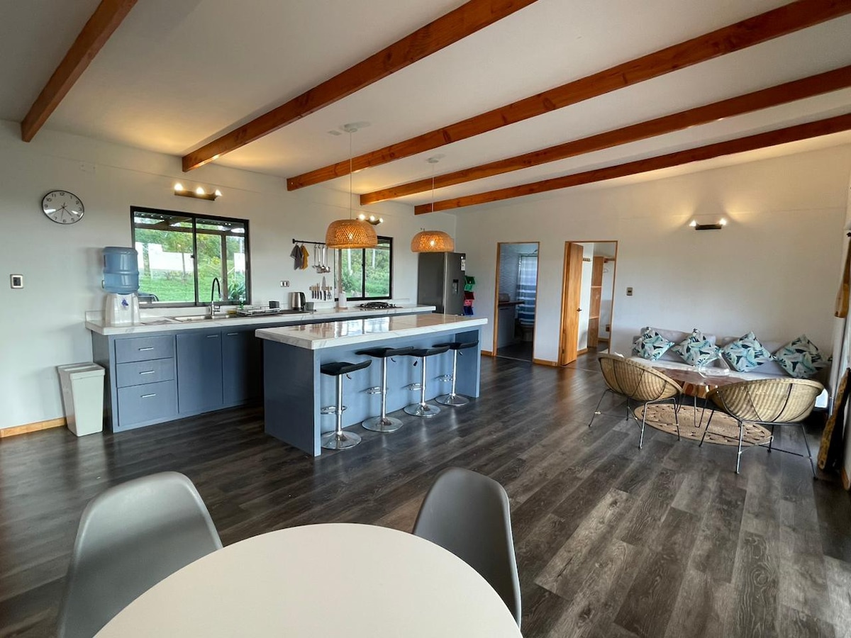 A spacious kitchen and dining area features a large island with seating for four. Modern light fixtures hang above. A dining table is positioned nearby, while cozy seating made of woven material adds comfort. Large windows allow natural light to fill the room.