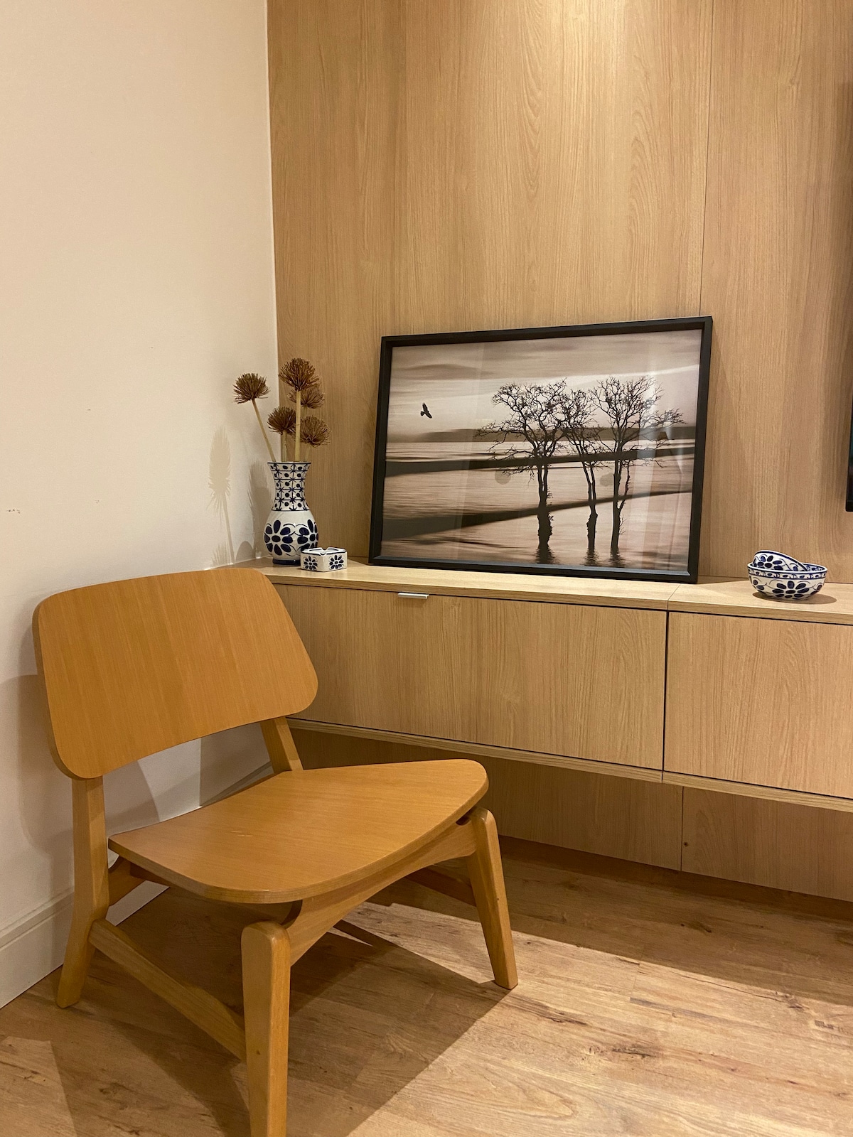 A wooden chair is positioned next to a light-colored wooden surface, complemented by a framed black-and-white photograph of trees along a body of water. Decorative items, including a floral arrangement and a patterned vase, add visual interest to the space.