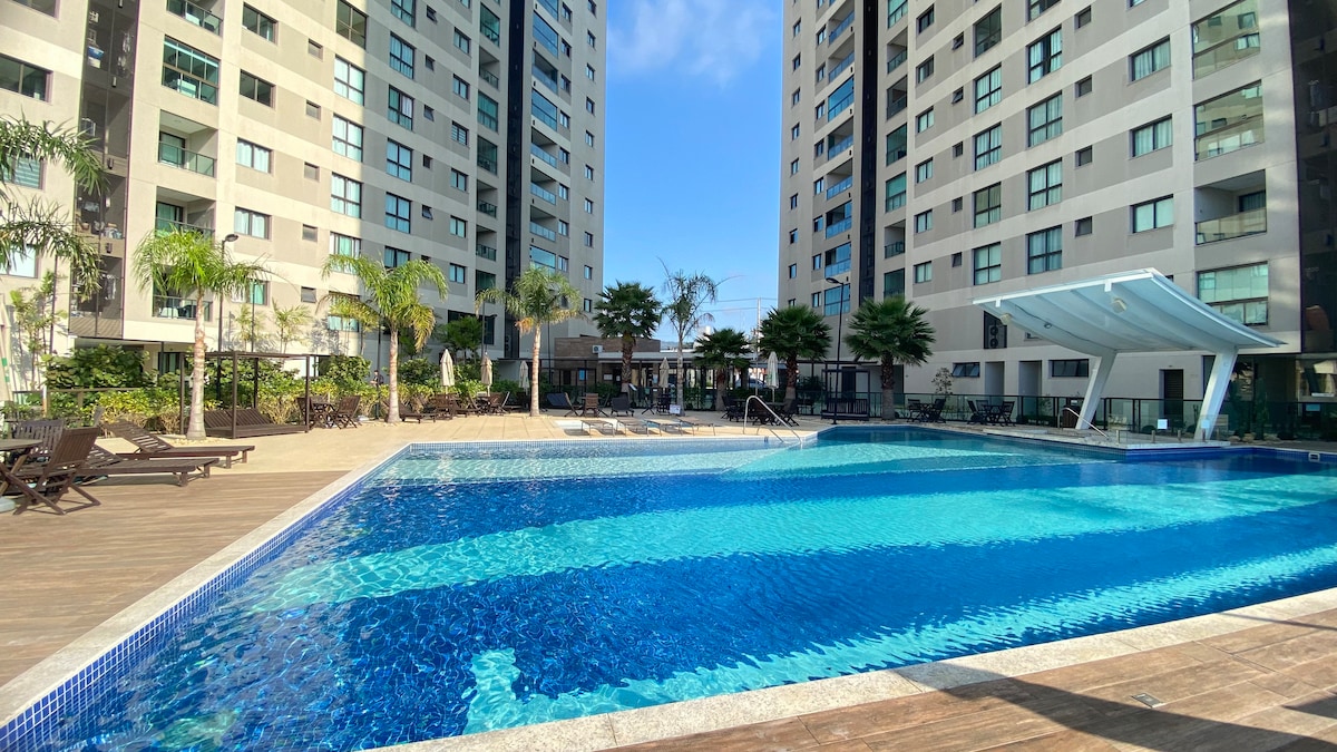 The image features a large swimming pool surrounded by tall palm trees and modern apartment buildings. Sun loungers line the wooden deck, and a shaded area is visible near the pool for relaxation. Clear blue skies reflect in the pool's inviting water.