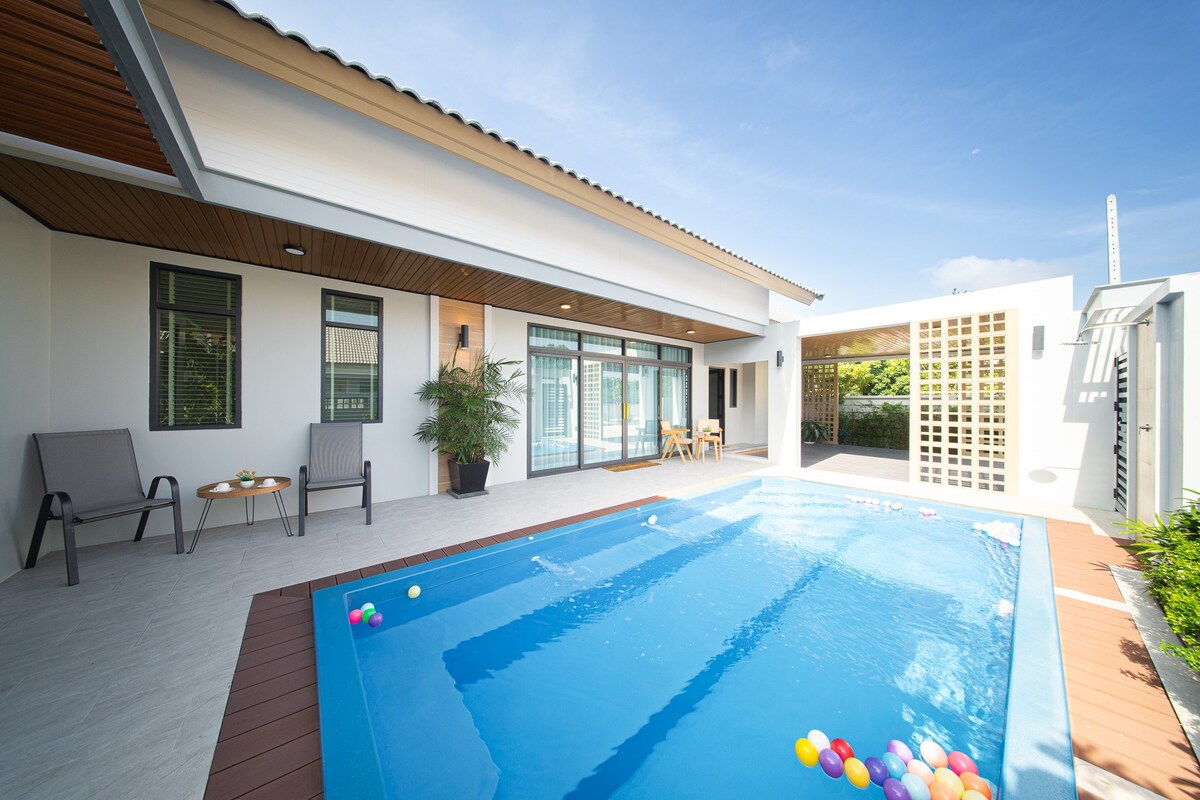 A private pool area is illuminated by natural light, featuring a clear blue pool surrounded by colorful floating balls. Two chairs and a small table are positioned on the patio, while large glass doors provide views into the interior space.