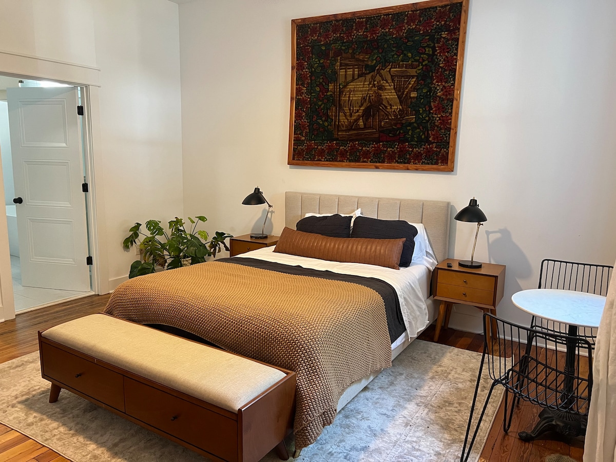A spacious bedroom is presented with a comfortable bed adorned with layered bedding. A decorative tapestry hangs above the bed, while two bedside lamps provide soft lighting. A wooden bench sits at the foot of the bed, accompanied by a small round table and a chair.