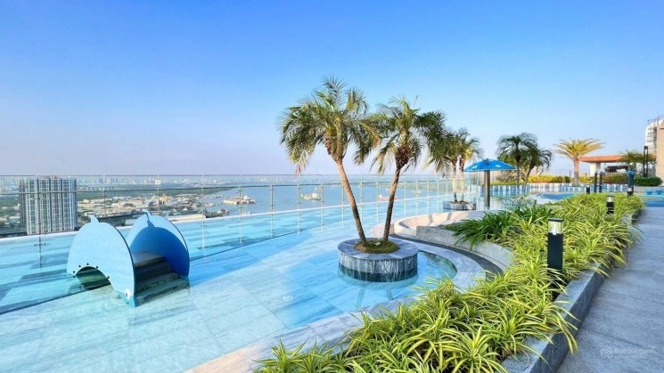 A spacious rooftop pool area is surrounded by well-maintained greenery and palm trees. Comfortable seating areas and umbrellas provide shade, while panoramic views of the river and city skyline create a serene backdrop for relaxation.