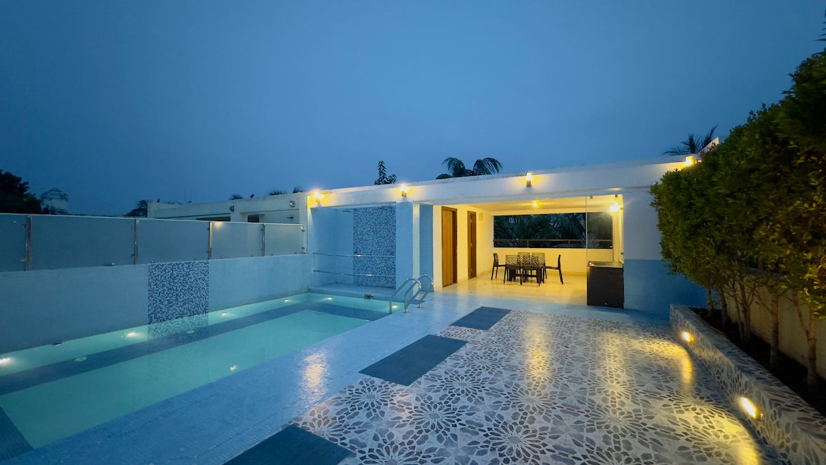 A tranquil pool area is presented, with water illuminated by subtle lighting. Surrounding greenery provides a natural border. The outdoor patio features a dining set, offering a welcoming space for relaxation. The structure showcases an open design, enhancing the evening ambiance.