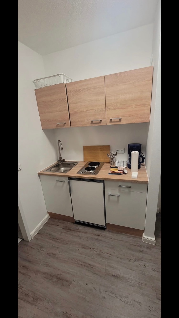 A compact kitchen area is featured with light wood cabinetry and a stainless-steel sink. A small countertop includes a cutting board and coffee maker, while a dishwashing unit is positioned beneath a counter. Neutral tones and a wooden floor create a cohesive and functional space.