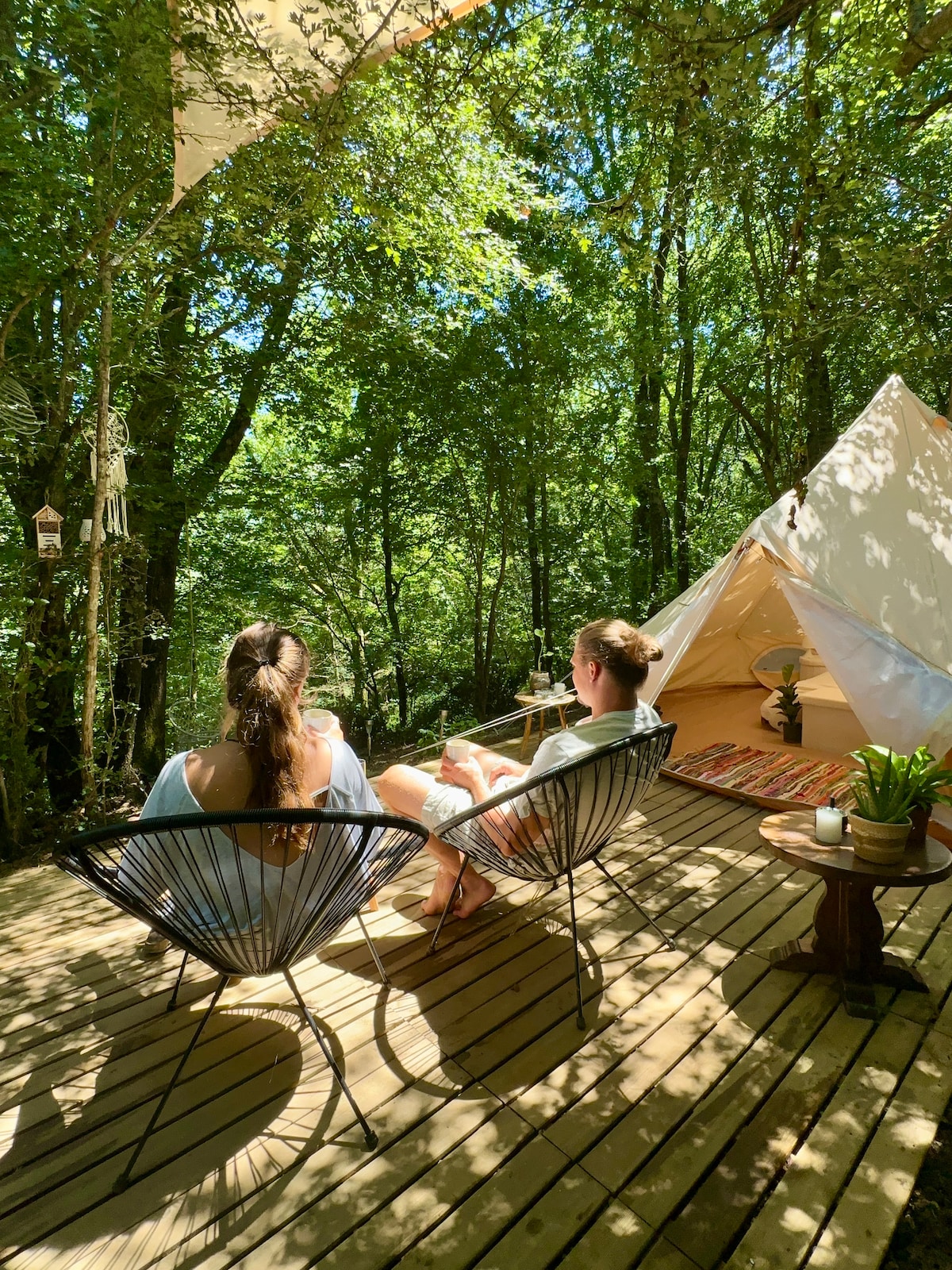 Two people are seated on black woven chairs on a wooden deck surrounded by lush green trees. A beige tent is visible in the background, with a colorful rug placed on the ground and a small table holding a potted plant nearby. Natural sunlight filters through the leaves.