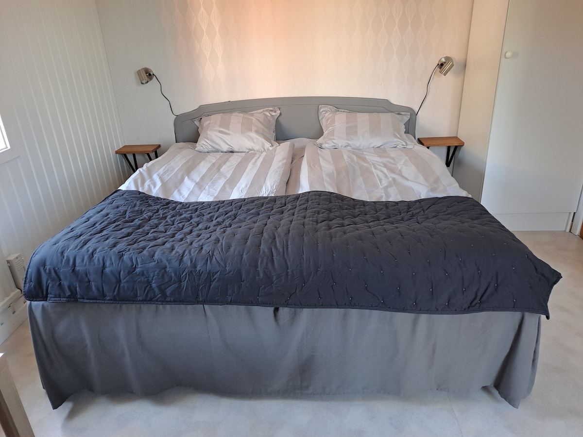 A spacious bed is featured with a dark blue quilt and two light-colored pillows. Bedside tables are positioned on either side, with wall-mounted lamps providing lighting. The room's light-colored walls add to the overall bright atmosphere.