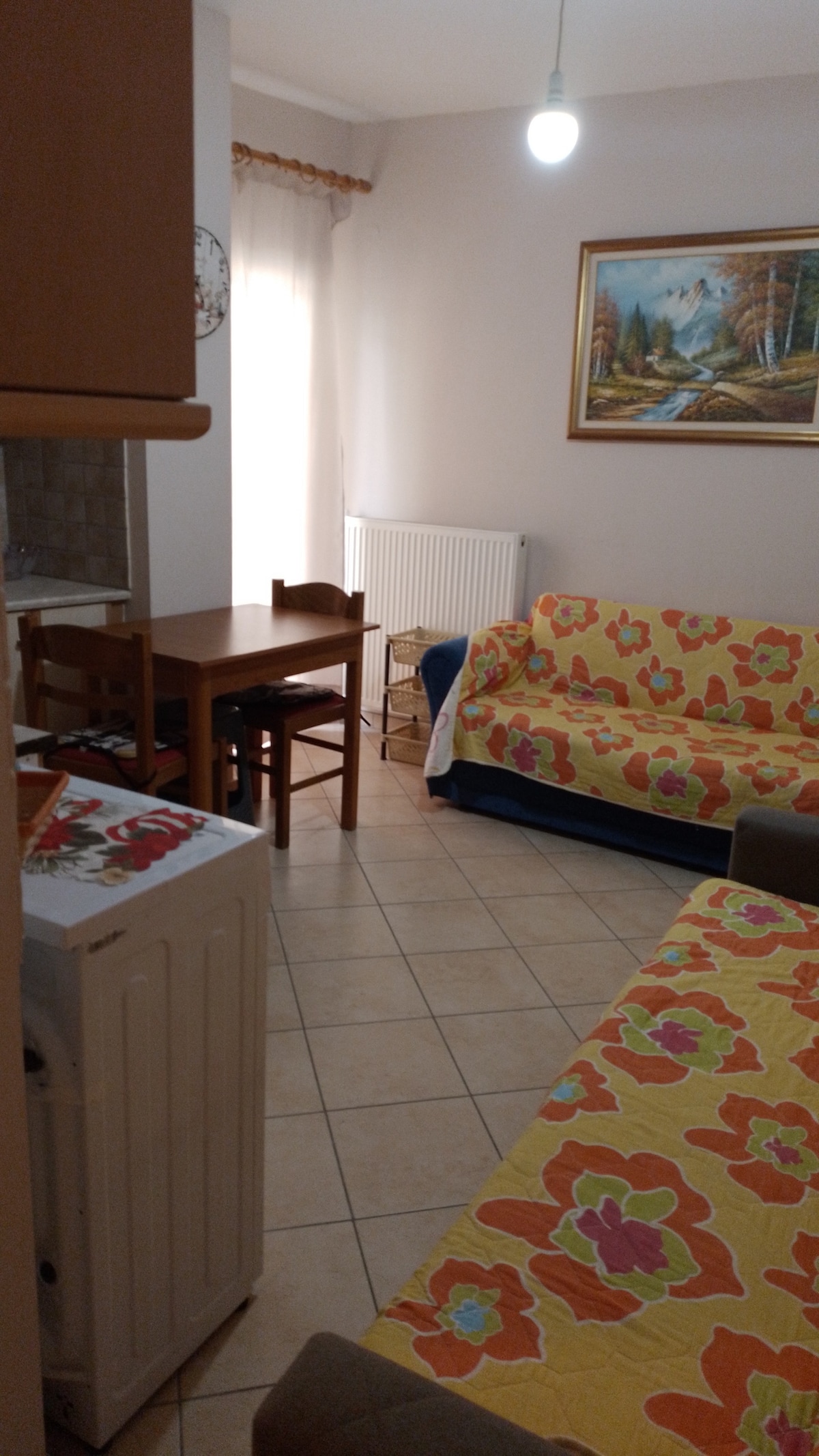A cozy living area features two sofas adorned with vibrant floral covers. A wooden dining table with chairs is positioned nearby, along with a washing machine. Natural light enters through a window, illuminating the warm-toned tiled floor.