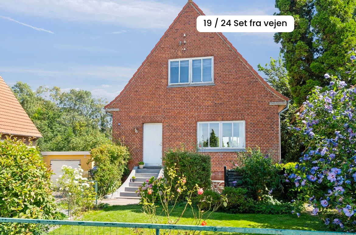 A charming brick house with a gable roof is surrounded by vibrant greenery and colorful flowers. A set of stairs leads to the front door, while large windows allow natural light indoors. The well-maintained garden offers a welcoming entrance.