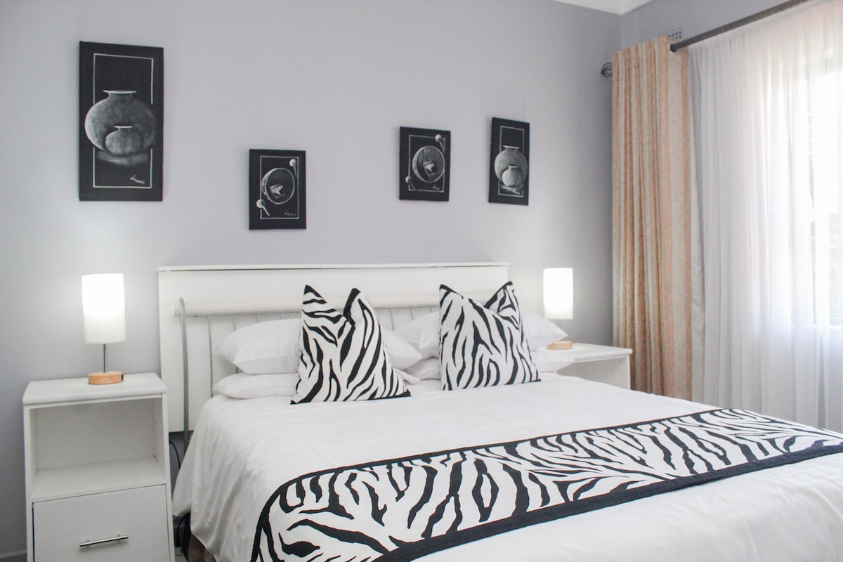 A well-appointed bedroom features a spacious bed adorned with patterned throw pillows. Two bedside lamps provide soft lighting on either side. Dark-framed artwork is displayed above the bed, and light curtains gently filter natural light from the window.