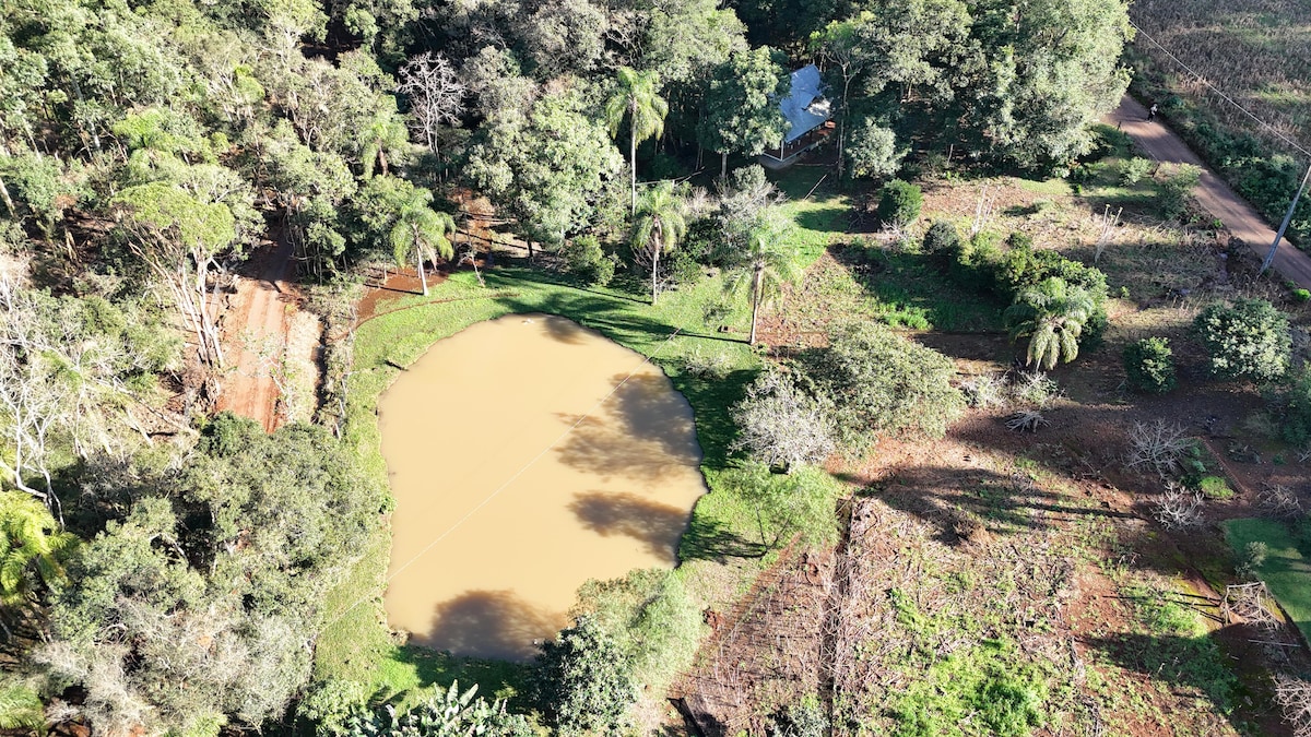 An aerial view captures a tranquil scene featuring a circular pond surrounded by lush greenery. Trees and scattered vegetation frame the area, while a small cabin is glimpsed in the background. The earth is rich with colors, reflecting a serene natural setting.