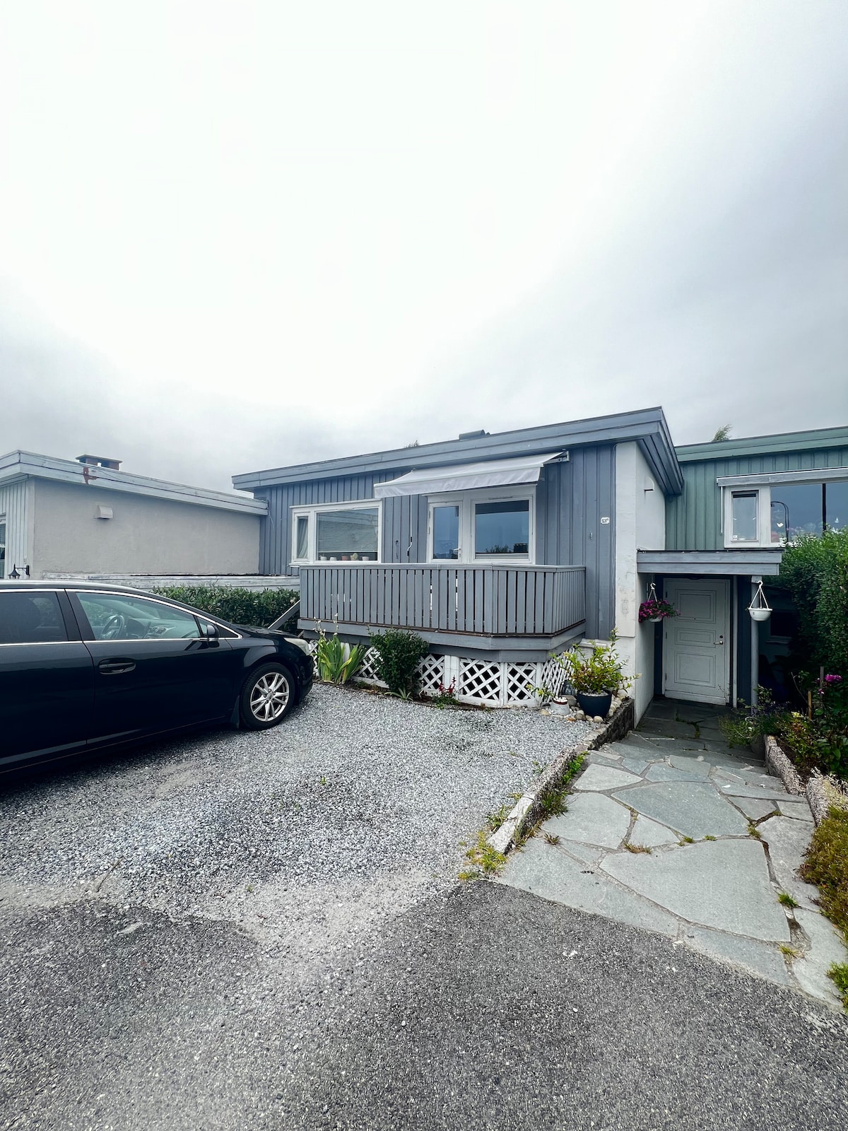 A modest house features a light gray exterior, with large windows allowing natural light to enter. A gravel driveway accommodates a black car, while decorative plants and a stone pathway lead to the entrance, enhancing the welcoming appearance of the home.
