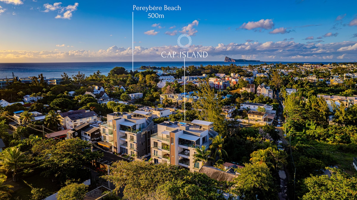 An aerial view highlights the penthouse's location within the Cap Island residence. The lush greenery surrounds the modern buildings, with Pereybere Beach visible in the distance, approximately 500 meters away. The clear sky and coastline create a serene backdrop.