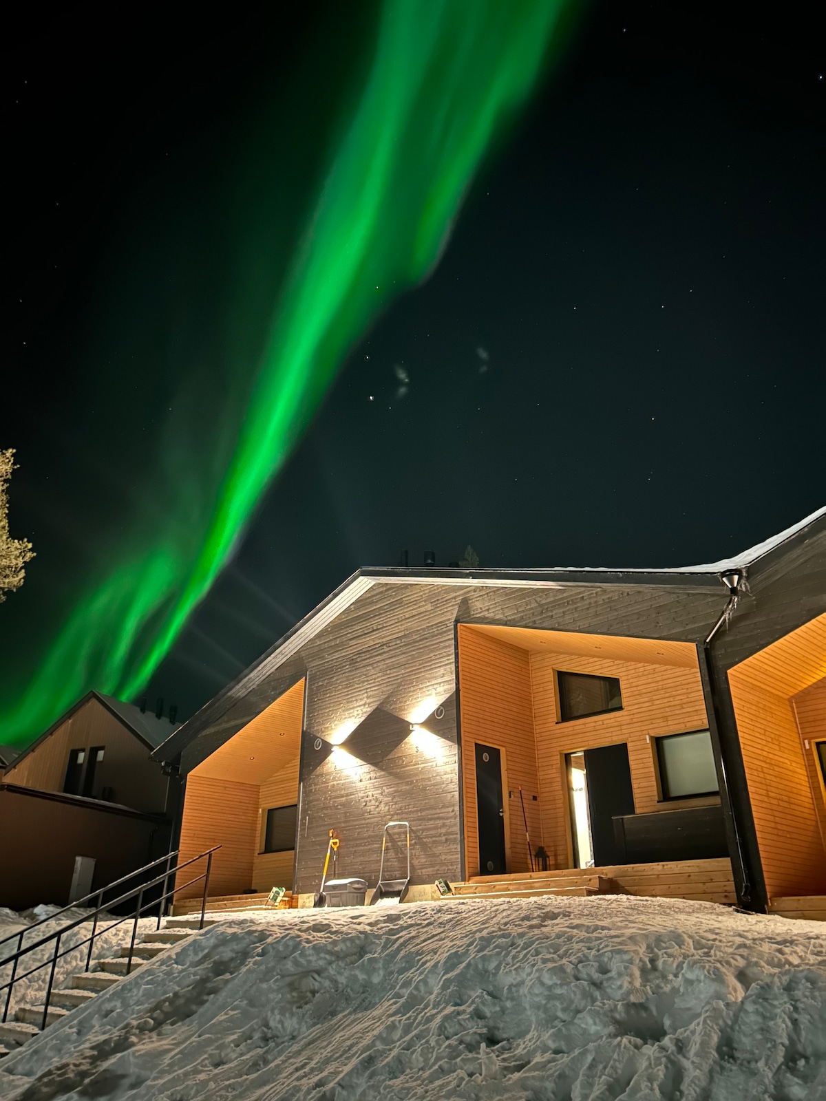 The exterior of a modern building is showcased against a night sky illuminated by the vibrant green hues of the northern lights. Snow covers the ground, and the structure features warm lighting that highlights its architectural details.