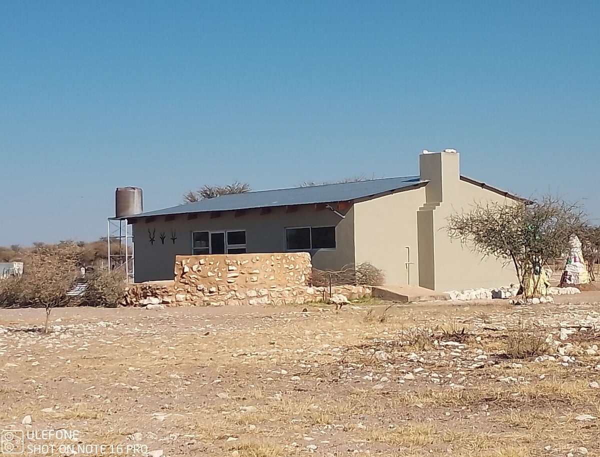 A single-story house is depicted with a sloped roof and weathered stone accents surrounding the entrance. The structure is set against a clear blue sky and is surrounded by sparse vegetation. A water tank is positioned on the roof, enhancing its rural charm.