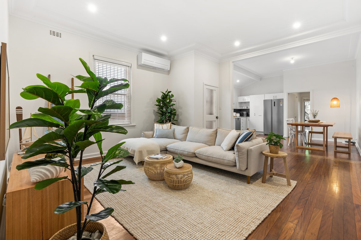 An open living area features a spacious, neutral-toned couch, surrounded by natural light from windows. Decorative plants are placed strategically, enhancing the room's serene atmosphere. The warm wooden flooring complements the area rug, while a dining space with a wooden table, chairs, and kitchen appliances is visible in the background.
