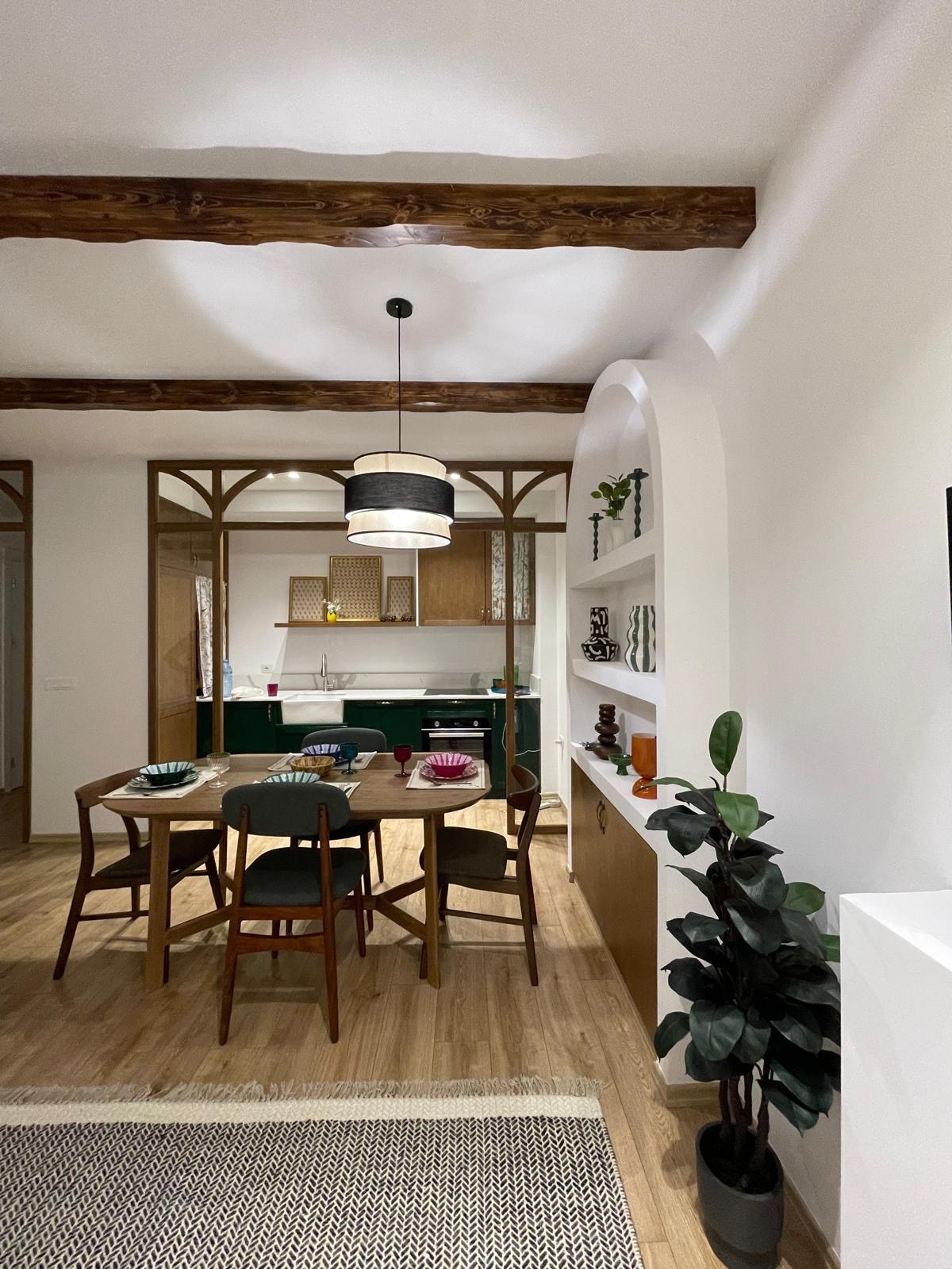 A dining area is depicted, featuring a round wooden table surrounded by four chairs. Light wood beams run across the ceiling, and the space is defined by white walls. Green cabinetry is visible in the background, complemented by neatly arranged shelves with decorative items.