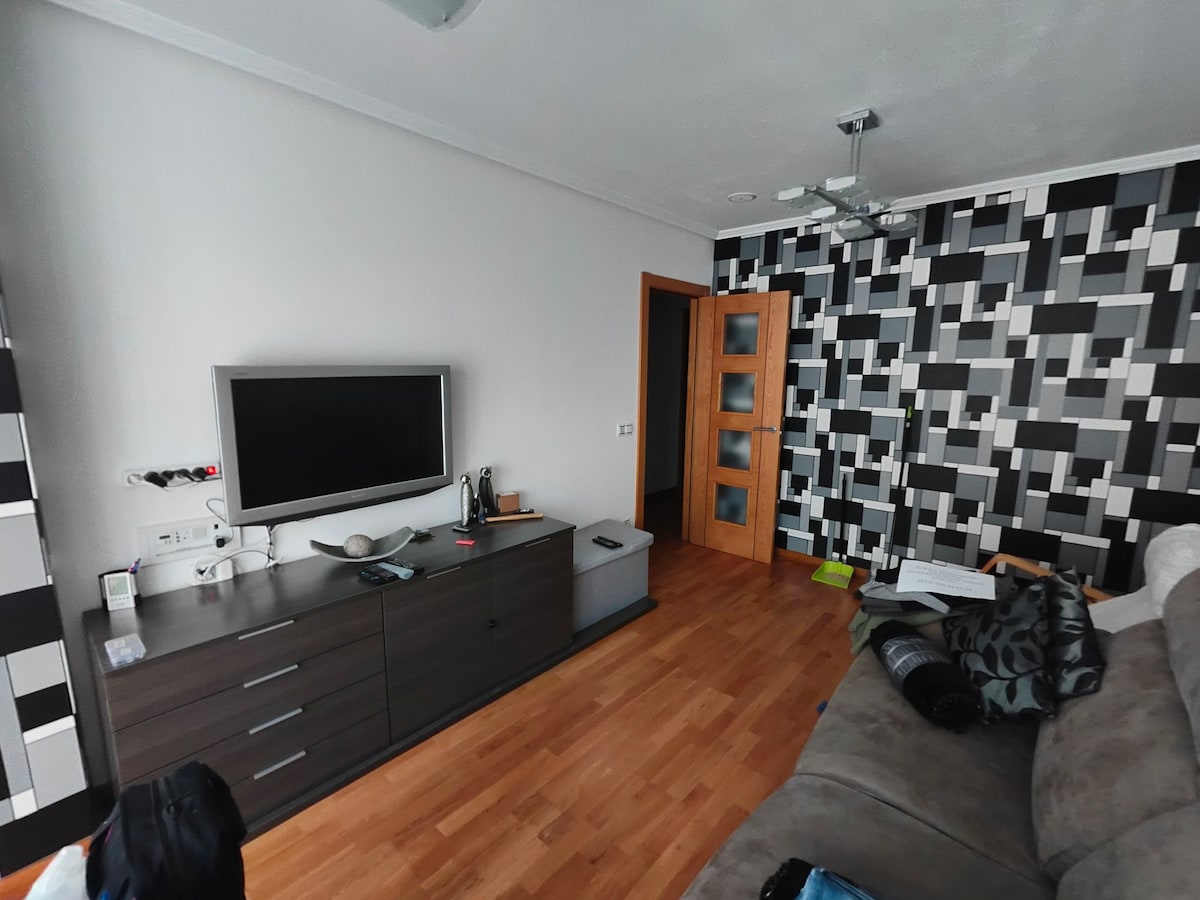 A spacious living area features a modern television mounted on a dark wood cabinet, complemented by a textured black and white accent wall. Light wood flooring adds warmth, while a doorway leads to another room. A comfortable sofa and decorative cushions are arranged for relaxation.