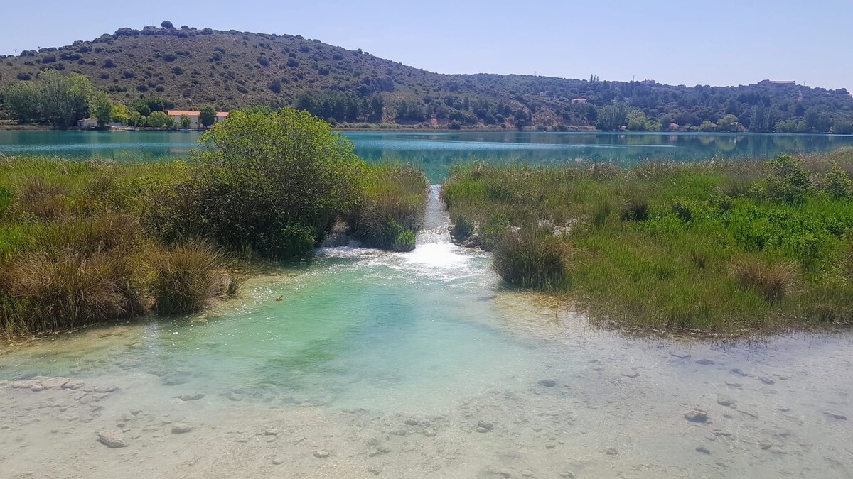 A serene landscape features a clear waterway flowing through lush green vegetation. The calm waters reflect the surrounding hills and plants, creating a peaceful scene. Sunlight illuminates the water's surface, enhancing the clarity of the shallow areas.