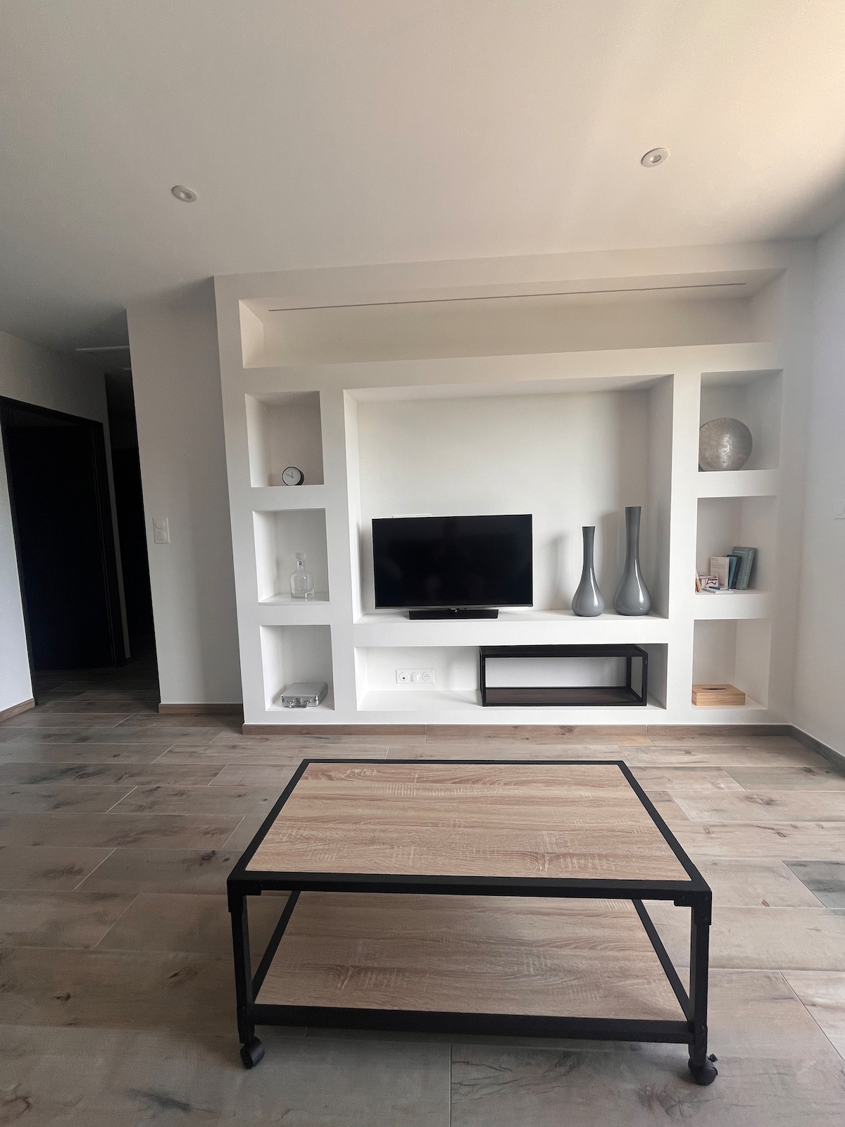 A modern living area is featured, centered around a stylish coffee table made of wood and metal. Built-in shelves display decorative items, and a flat-screen TV is mounted on the wall. Soft natural light fills the space through nearby windows.