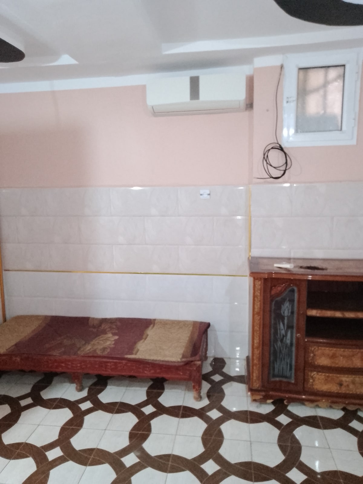 A simple room features a single wooden bed with a patterned blanket. A low wooden cabinet is positioned against the wall, and an air conditioning unit is mounted above. The room's walls are partially tiled, and the floor exhibits a unique geometric pattern.