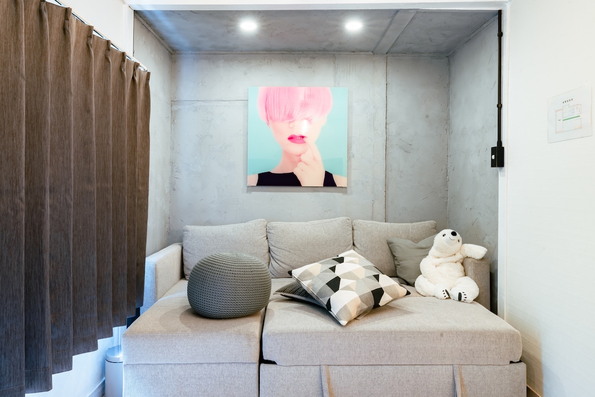 A cozy seating area features a light gray sectional sofa adorned with patterned cushions. Soft lighting illuminates the space, which includes a decorative round pillow and a plush toy. A colorful artwork hangs on the wall, while a textured curtain offers privacy.