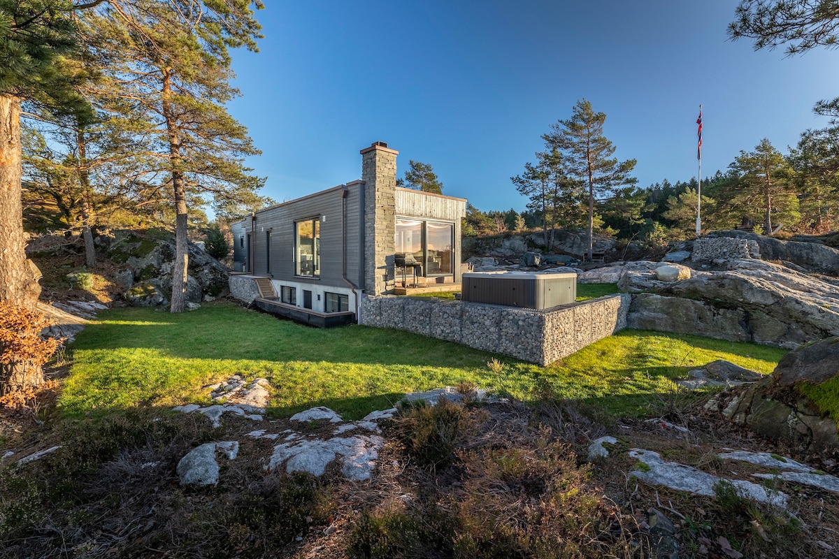 A modern cabin, constructed with large glass windows, is nestled among rocks and trees. A jacuzzi is positioned outside on a stone patio, surrounded by a well-maintained green lawn. The serene landscape offers a sense of privacy and tranquility.