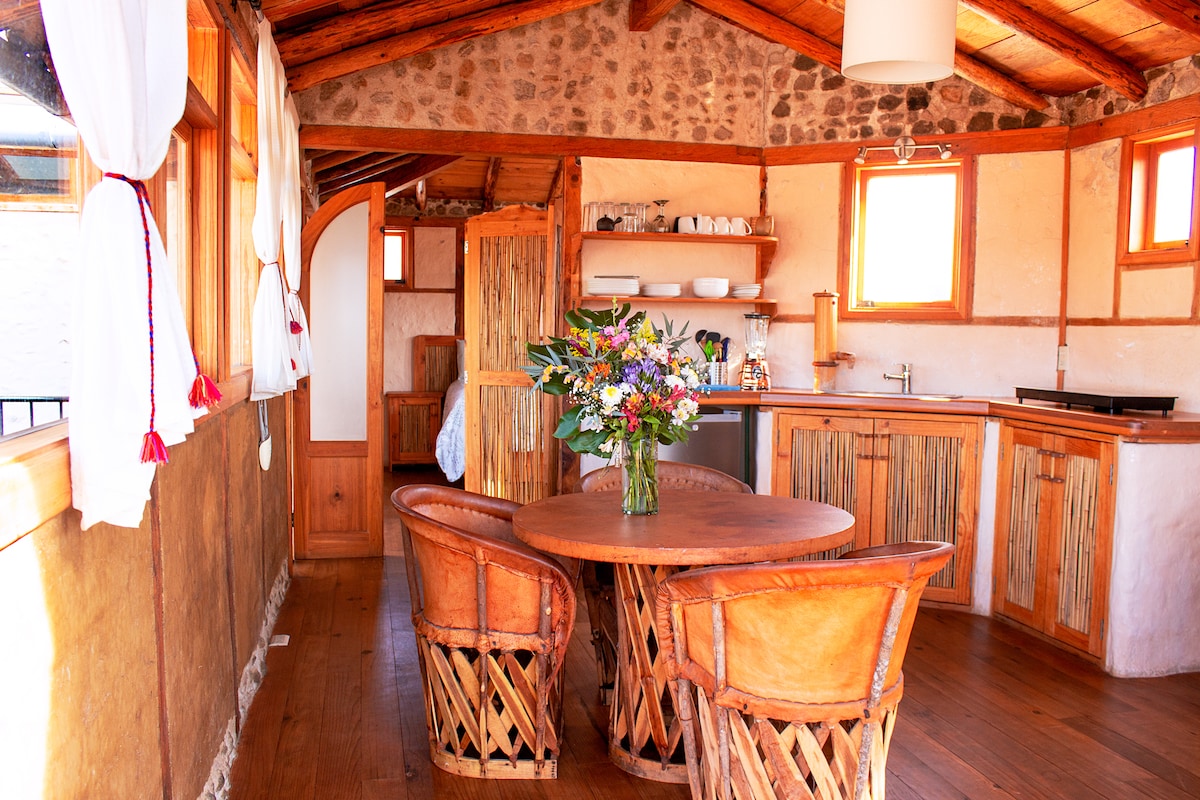 The kitchen area features wooden cabinetry and open shelving displaying dishware. A circular wooden dining table is surrounded by four woven leather chairs. Natural light enters through large windows adorned with white curtains, illuminating the warm tones of the rustic stone and wood interior.