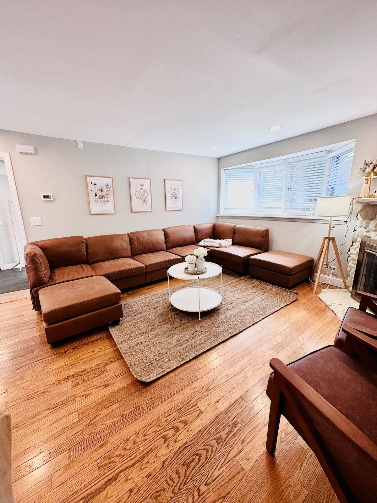 A spacious living area features a large brown sectional couch and a round coffee table. Sunlight filters through the windows, illuminating the hardwood floor. Wall art adorns the walls, complemented by a soft area rug and decorative floor lamp.